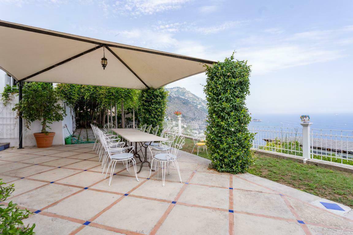 Geräumige Terrasse mit großem Esstisch unter einer Überdachung, umgeben von Grün und mit Meerblick.