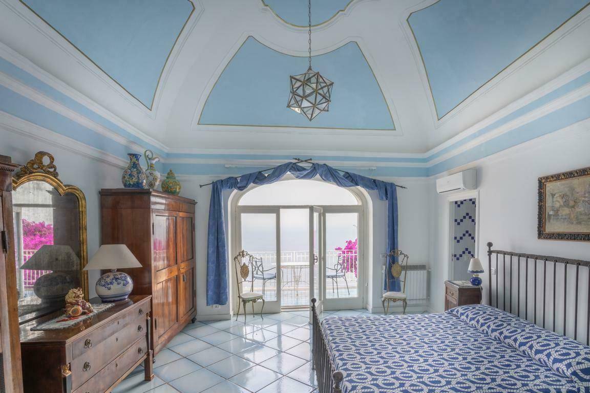 Elegantes Schlafzimmer mit hohen Decken, blauen Akzenten und Zugang zu einem französischen Balkon mit Meerblick.