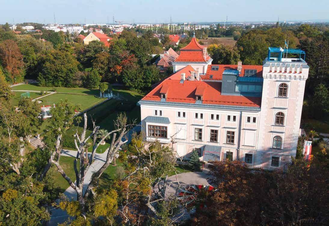 Luftaufnahme des historischen Gebäudes mit weitläufigem Park und städtischer Umgebung im Hintergrund.