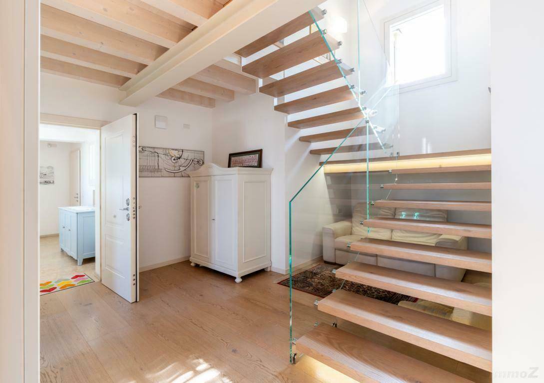 Heller Flur mit moderner Holztreppe, Glasgeländer und sichtbaren Deckenbalken.