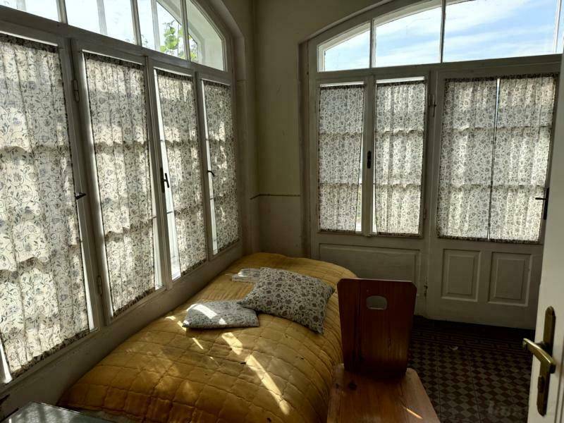 Helles Schlafzimmer mit vielen hohen Fenstern, floralen Vorhängen und gemustertem Fliesenboden.