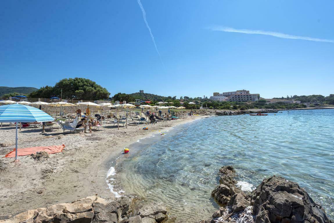 Belebter Sandstrand mit Sonnenschirmen und Liegen, umgeben von klarem Meerwasser und Küstenlandschaft.