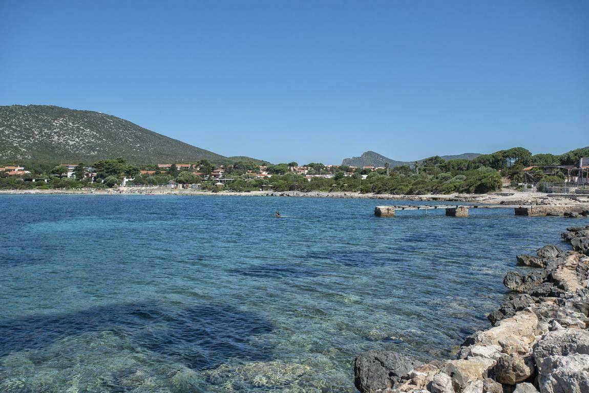 Klares, türkisfarbenes Meerwasser in einer ruhigen Bucht mit felsiger Küstenlinie und Bergen im Hintergrund.