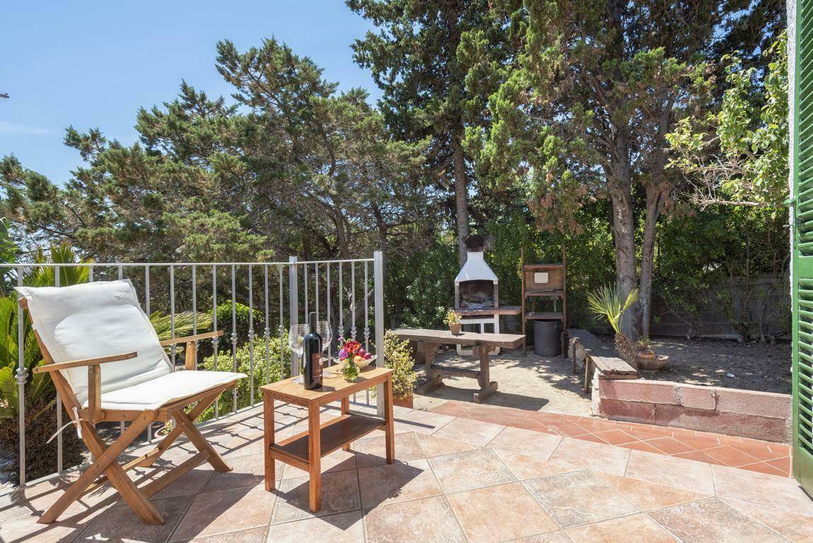 Sonnige Terrasse mit Grillbereich, Holztisch und Stühlen, umgeben von mediterraner Vegetation.