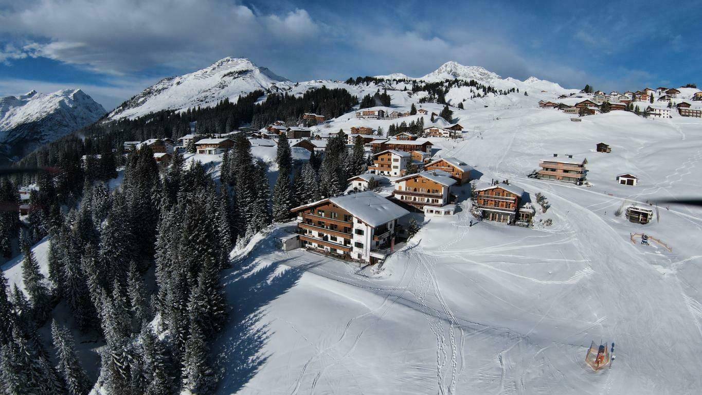 Luftaufnahme eines verschneiten Bergdorfes mit traditionellen Chalets, umgeben von Wäldern und majestätischen Bergen.