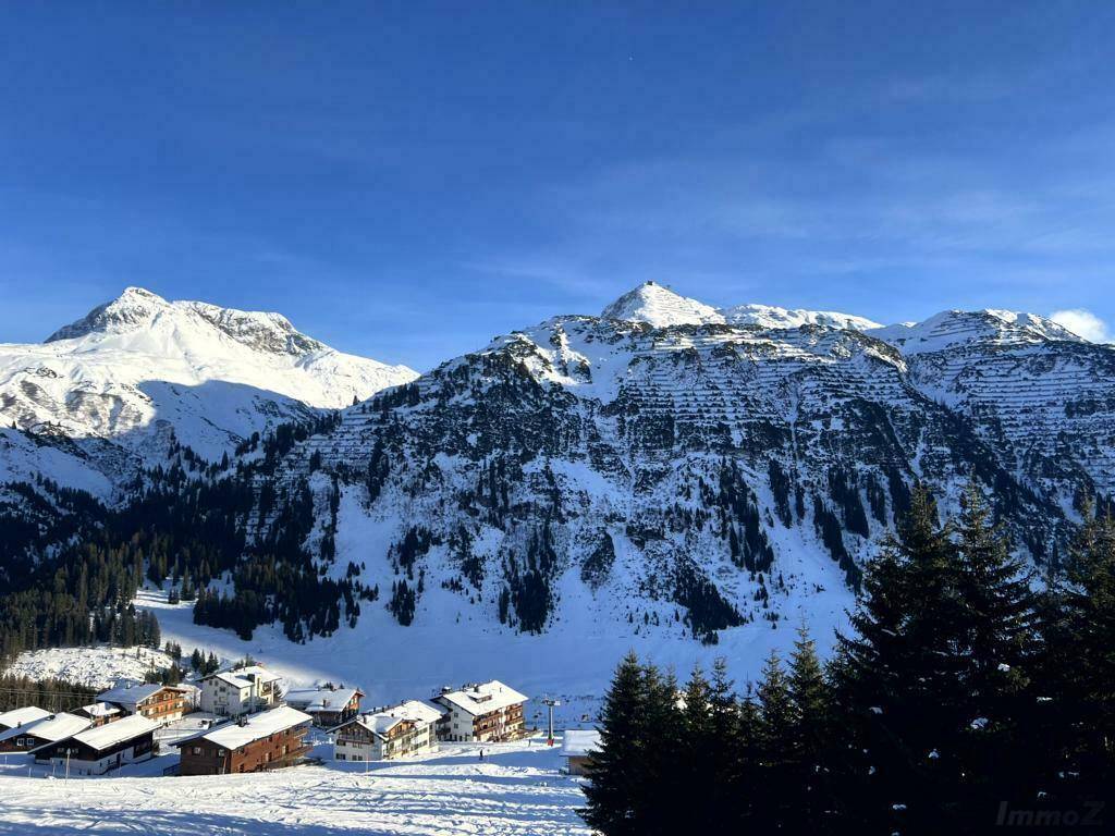 Panoramablick auf eine verschneite Berglandschaft mit einem kleinen Dorf im Tal unter blauem Himmel.