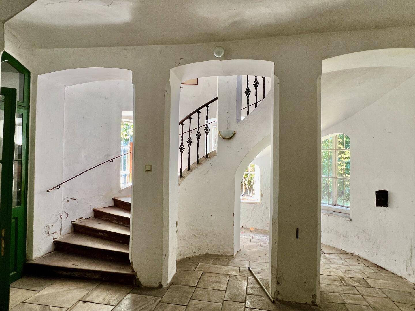 Historischer Flur mit geschwungenen Treppen, Rundbögen und Fliesenboden, mit viel Tageslicht.