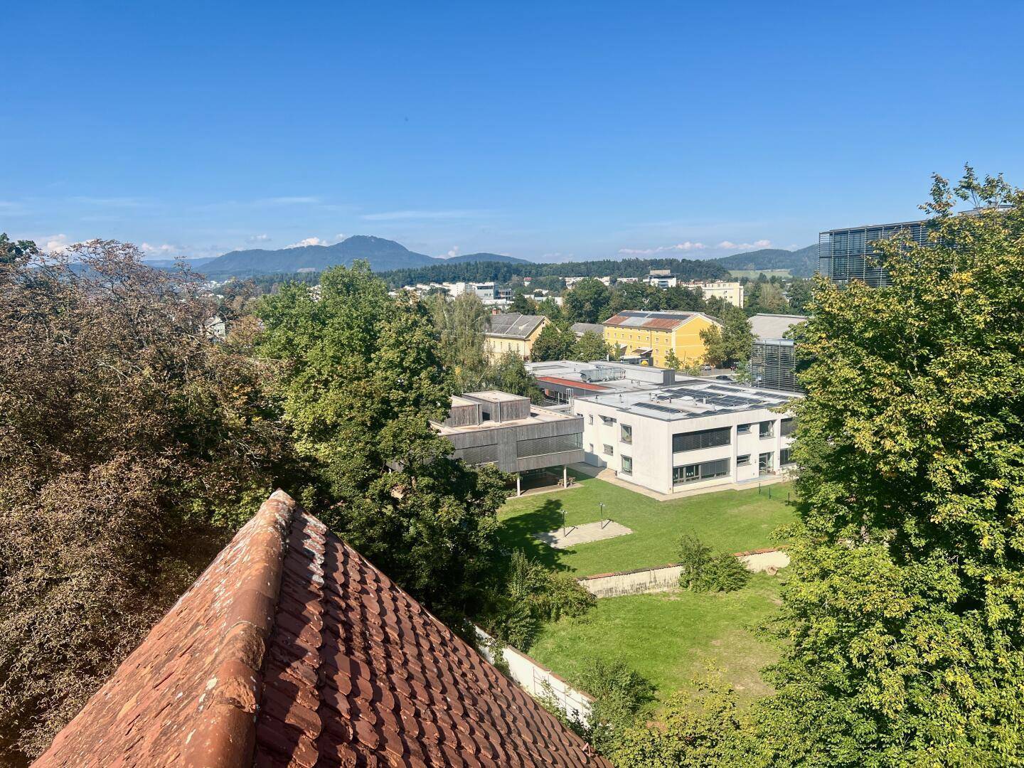 Weitblick über die Stadt und grüne Hügel bis zu den Bergen unter klarem Himmel.