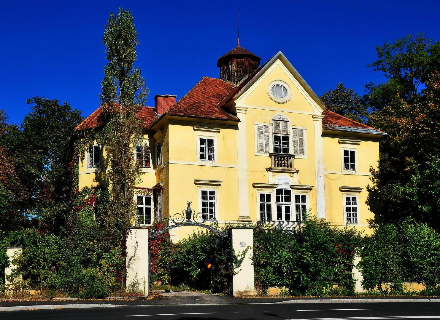 Historische Villa mit gelber Fassade, rotem Ziegeldach und schmiedeeisernem Tor, umgeben von Bäumen.