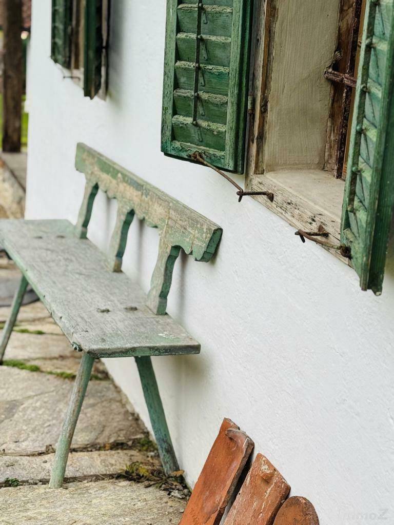 Alte Holzbank vor einer weiß getünchten Wand mit grünen Fensterläden und Steinboden.
