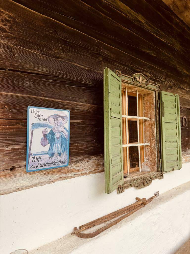 Rustikale Holzwand mit einem kleinen Fenster, grünen Fensterläden und einem traditionellen Schild.