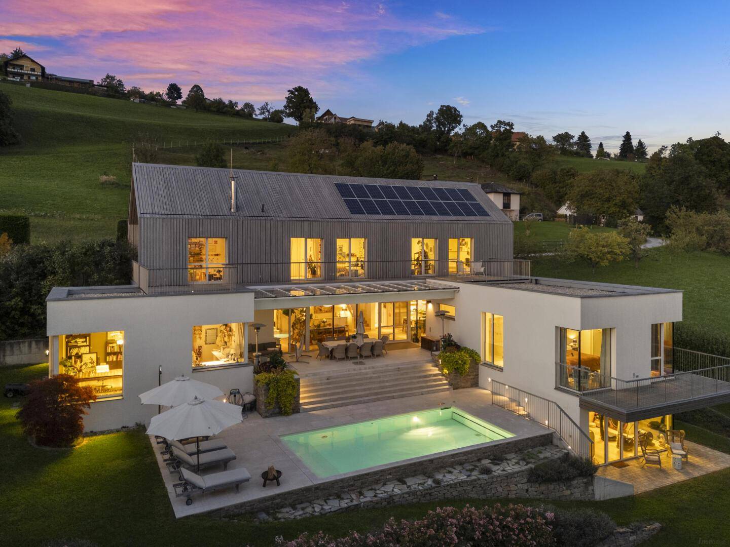 Moderne Villa mit beleuchtetem Pool und Terrasse in der Abenddämmerung, umgeben von Natur.