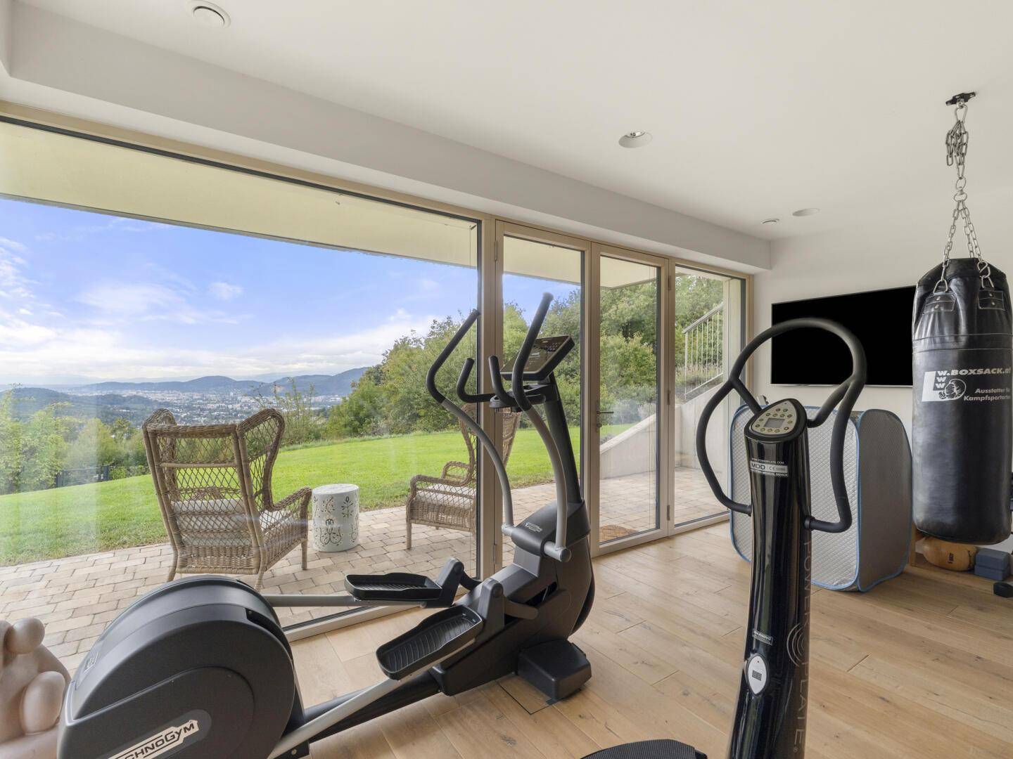 Heller Fitnessraum mit Trainingsgeräten, großen Fenstern und Blick ins Grüne.