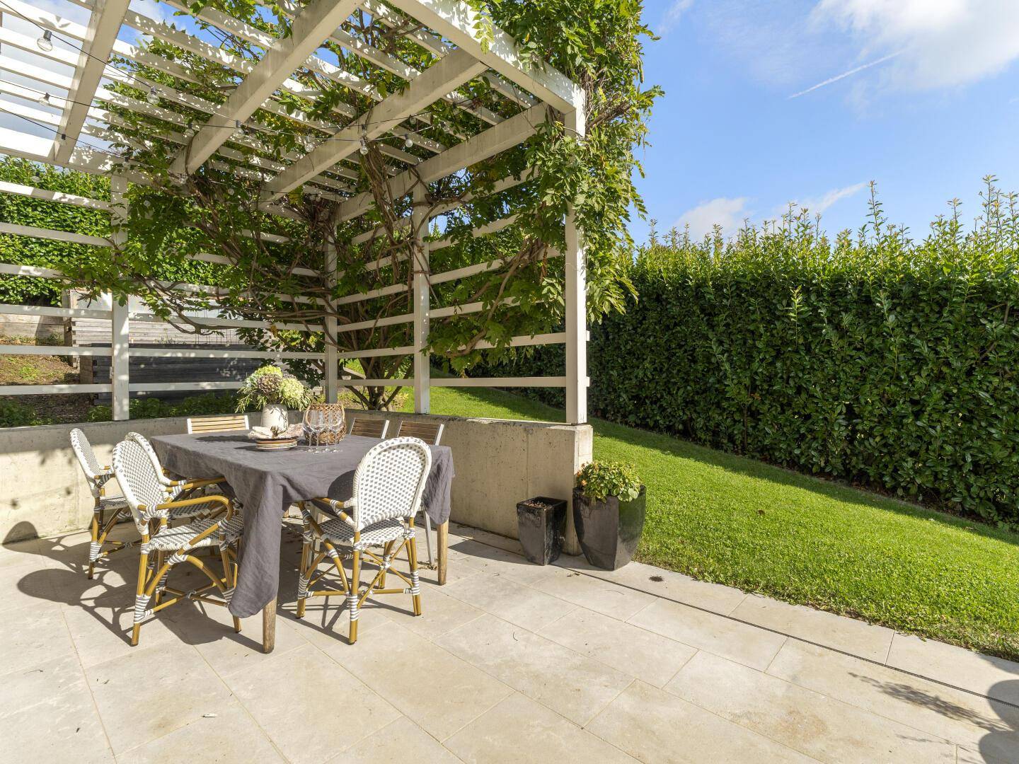 Gemütliche Gartenterrasse mit Esstisch unter einer bewachsenen Pergola und Hecke.