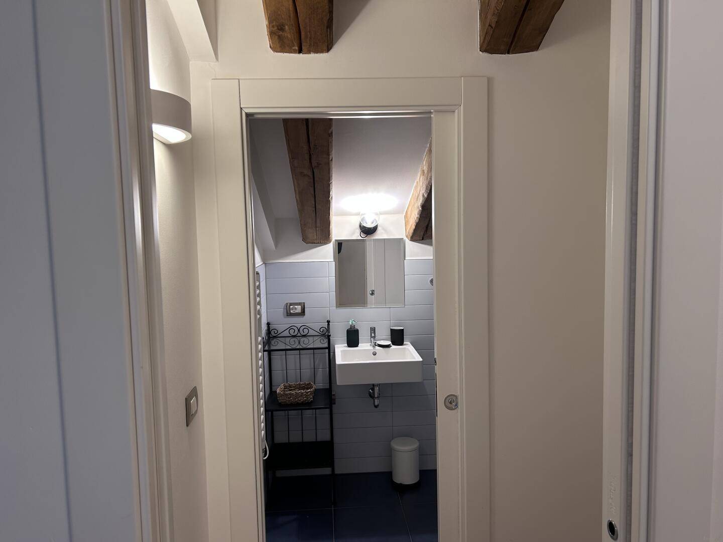 Blick durch die Tür in ein modernes Badezimmer mit hellblauen Fliesen und Waschbecken unter Holzbalken.