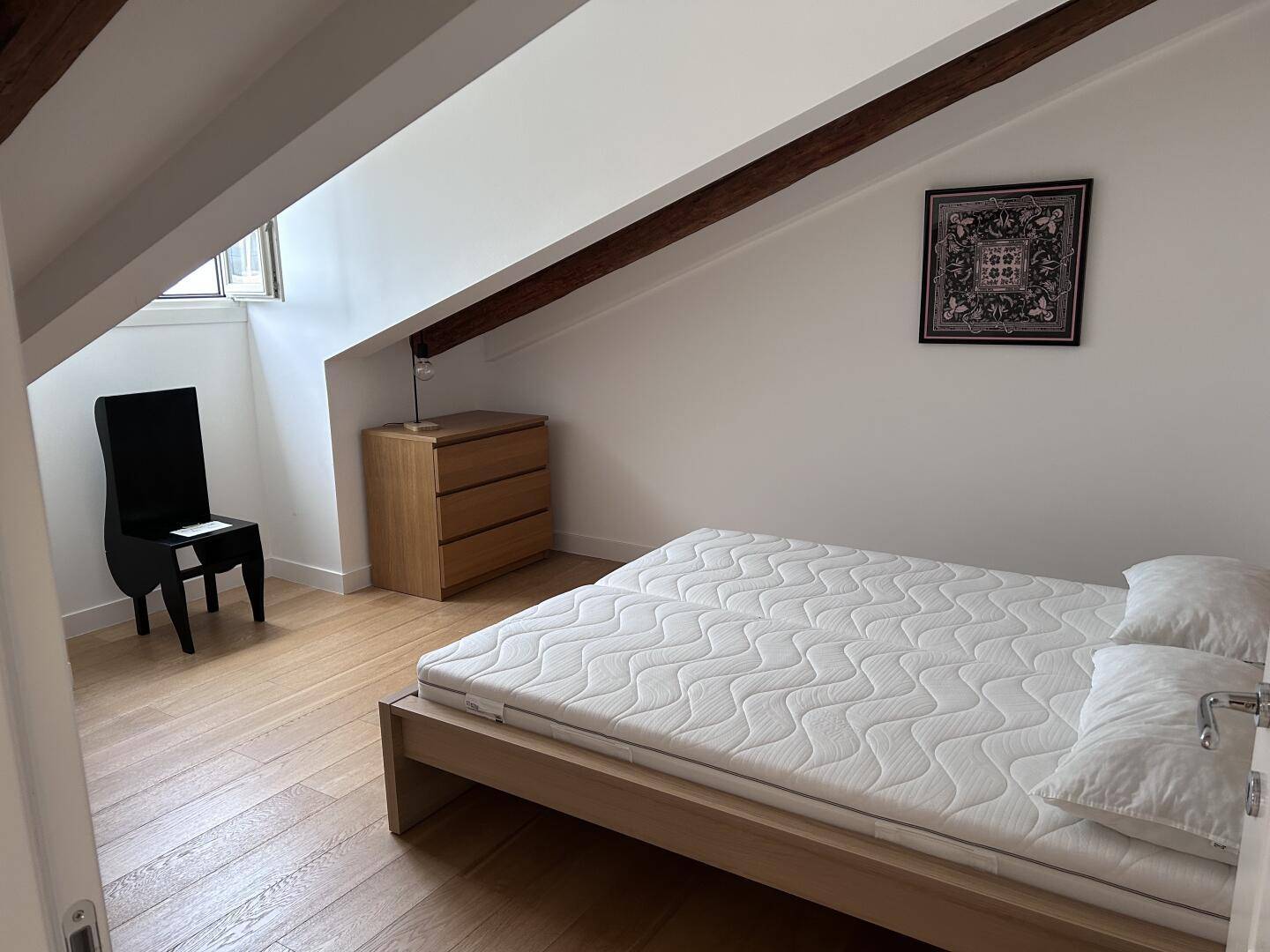 Helles Schlafzimmer mit Holzbalken, Parkettboden und einem einfachen Bettgestell unter dem Dach.