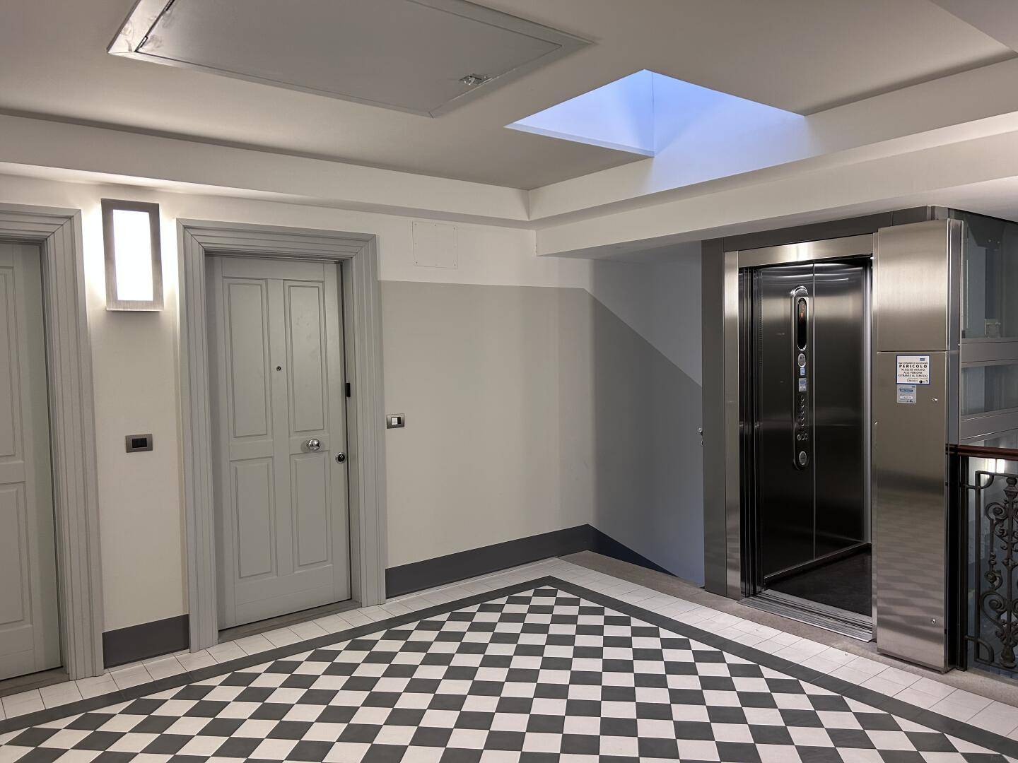 Heller Flur im Gebäude mit Schachbrettmuster-Bodenfliesen und einem modernen Aufzug.