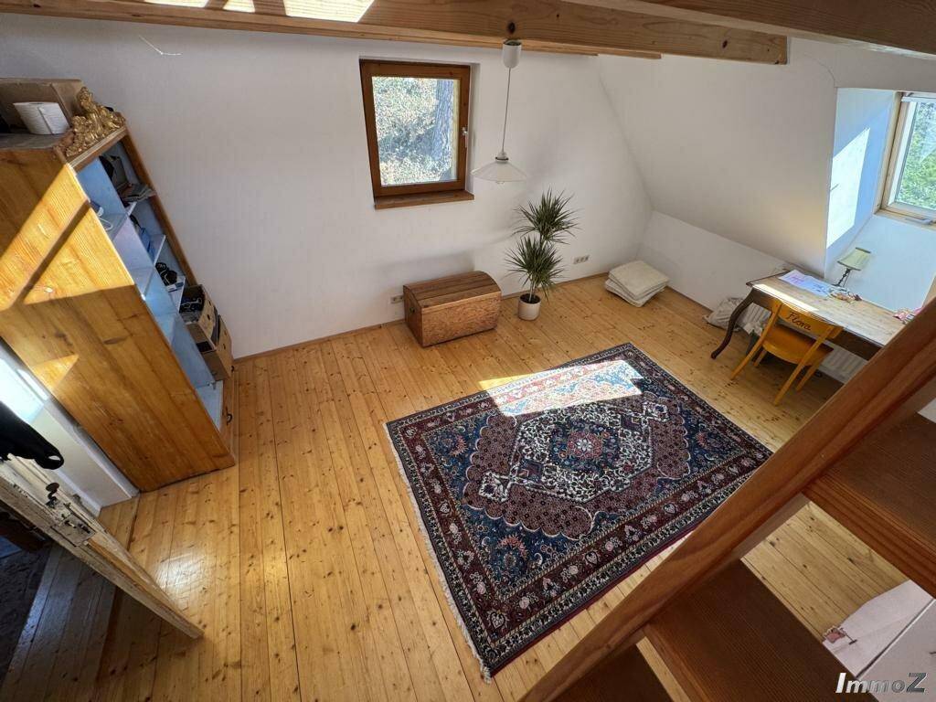 Helles Dachzimmer mit Holzdielenboden, sichtbaren Holzbalken und einem Fenster mit Blick ins Grüne.
