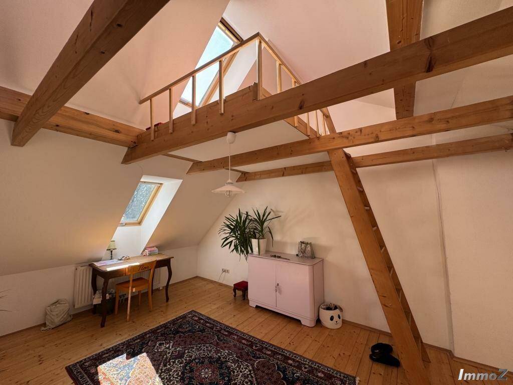 Geräumiges Dachzimmer mit Holzbalken, schrägen Wänden und einer Leiter zu einem oberen Bereich.