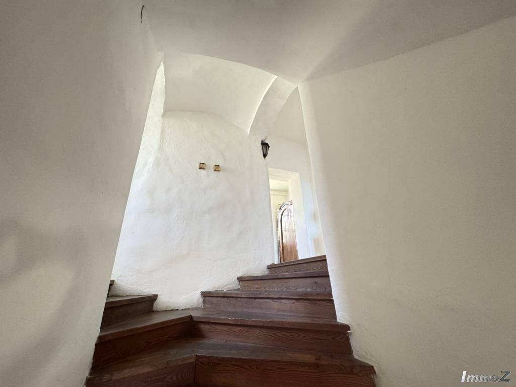 Unterer Bereich einer geschwungenen Holztreppe mit weiß verputzten Wänden und gewölbten Durchgängen.