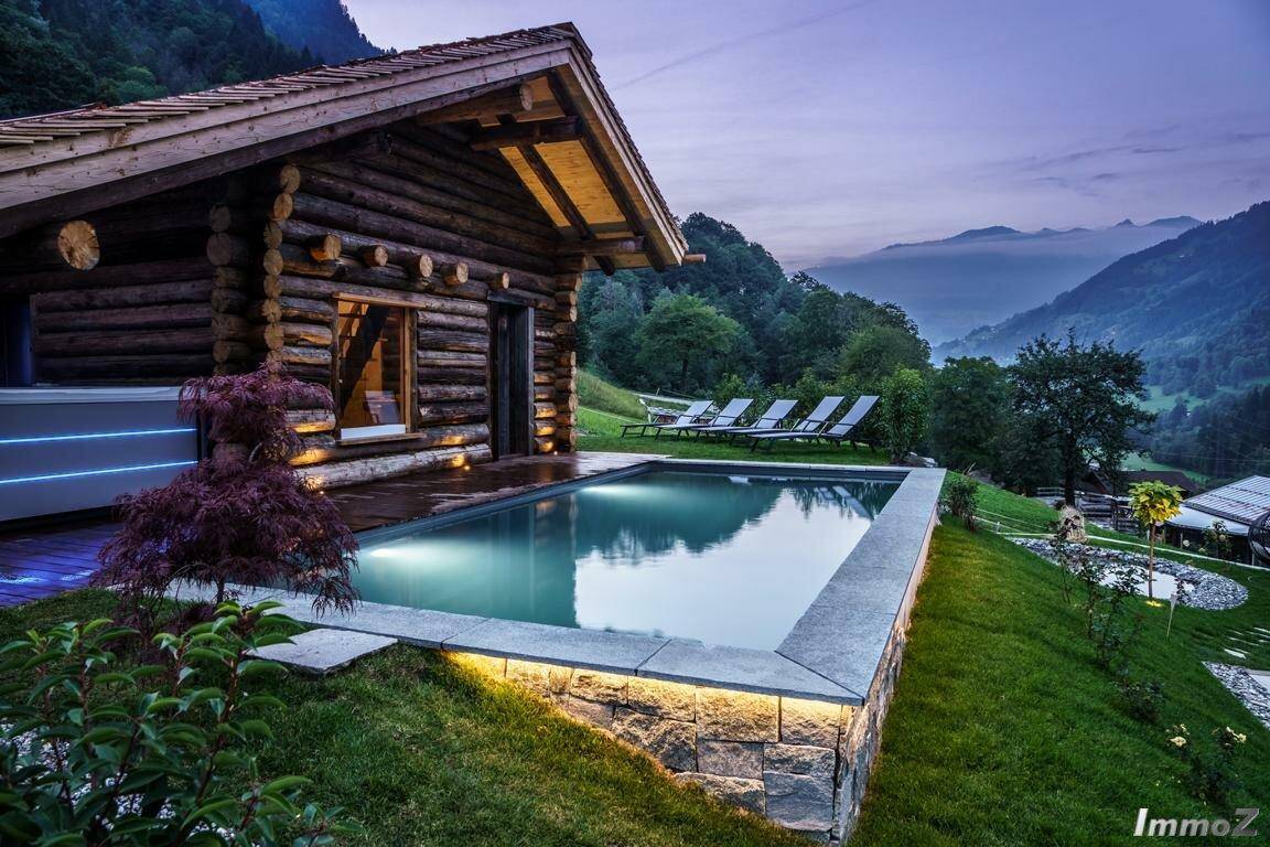 Beleuchteter Außenpool mit Bergblick und rustikaler Holzhütte in der Abenddämmerung.