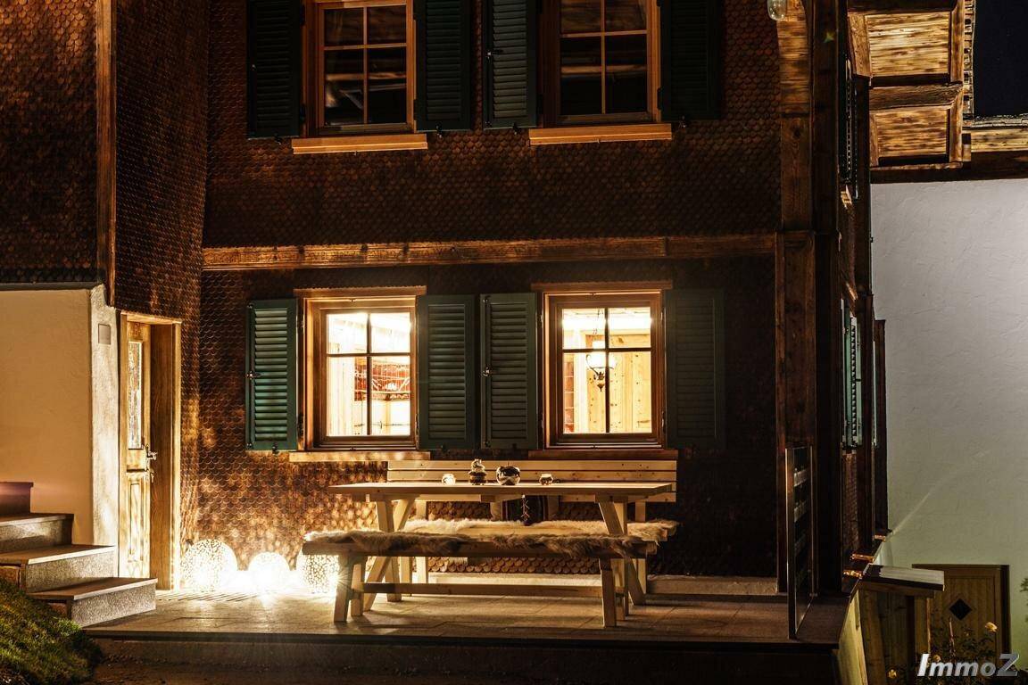 Rustikale Holzfassade eines Hauses bei Nacht mit beleuchtetem Essbereich im Freien.