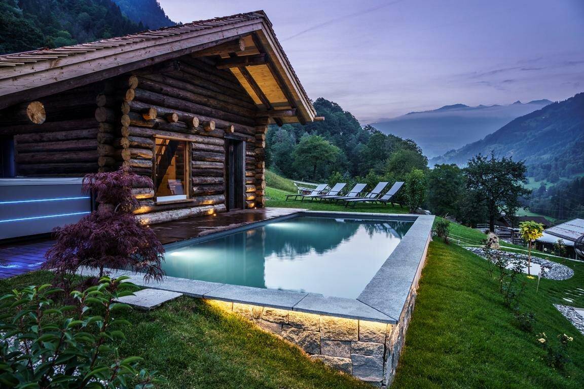 Beleuchteter Außenpool mit Whirlpool und Bergblick vor einer Holzhütte in der Dämmerung.