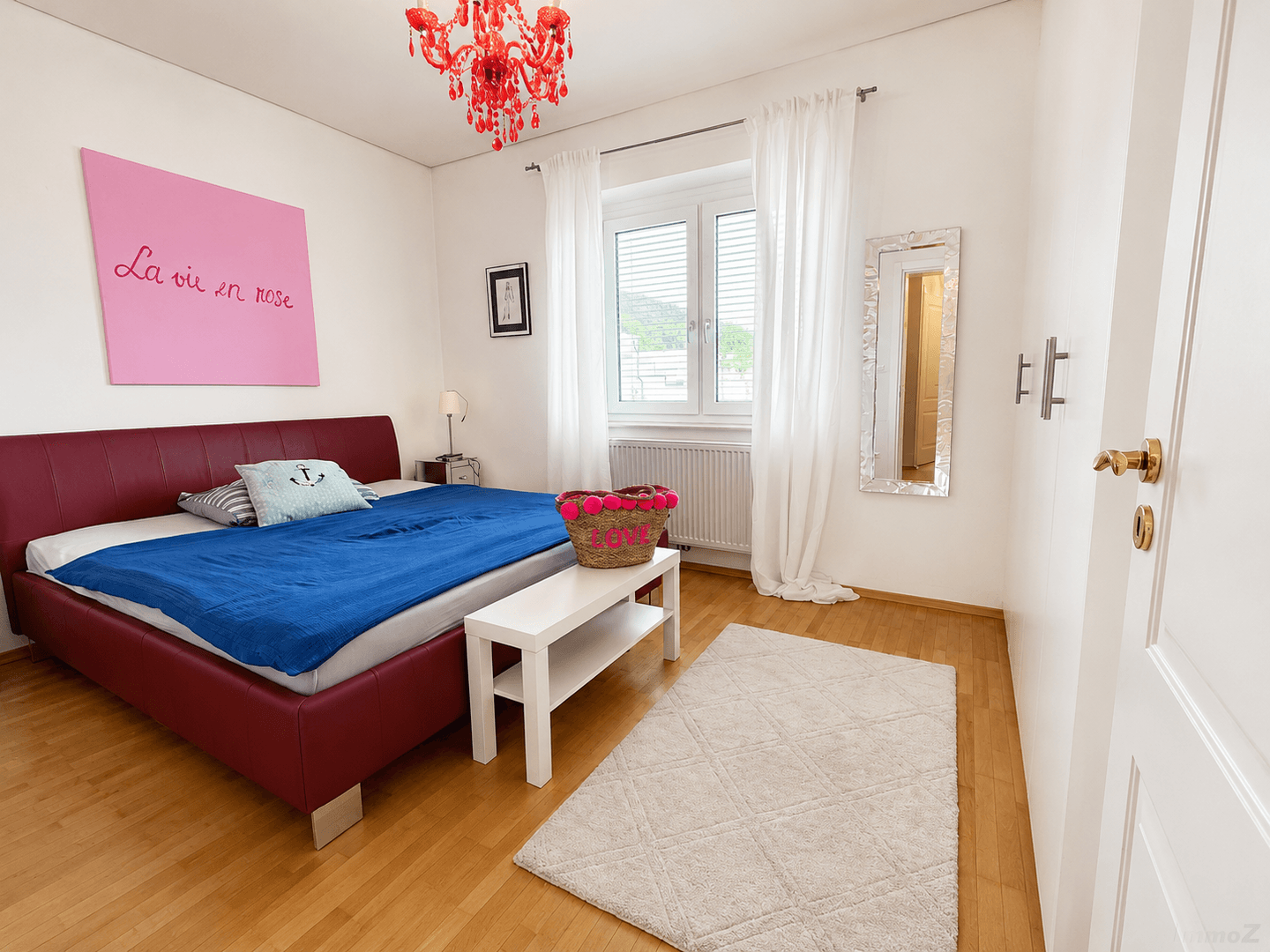 Schlafzimmer mit rotem Bett, Holzboden und großem Fenster für viel Tageslicht.