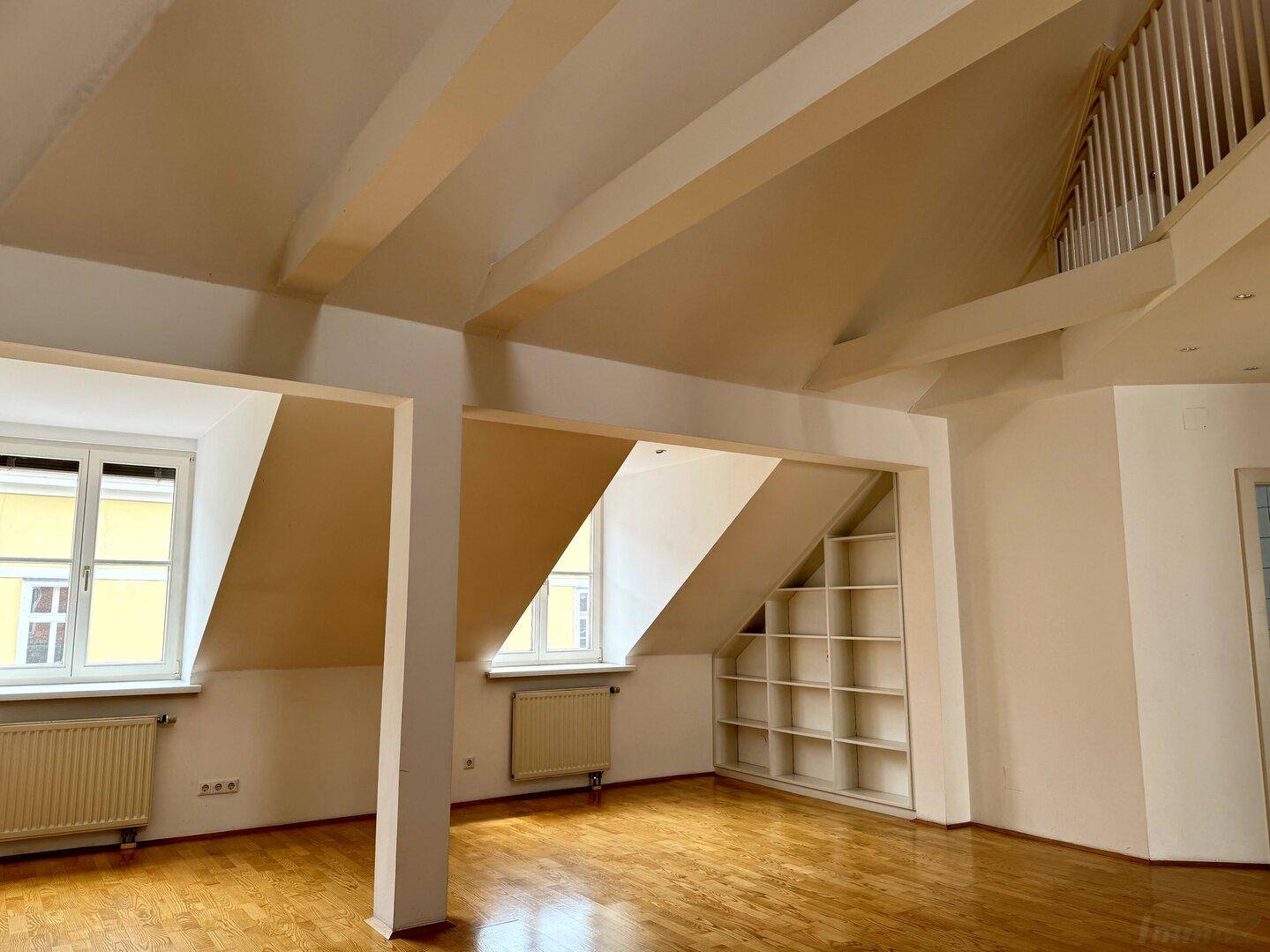 Heller Wohnbereich mit Holzboden, großen Fenstern, sichtbaren Balken und integriertem Regal unter der Dachschräge.