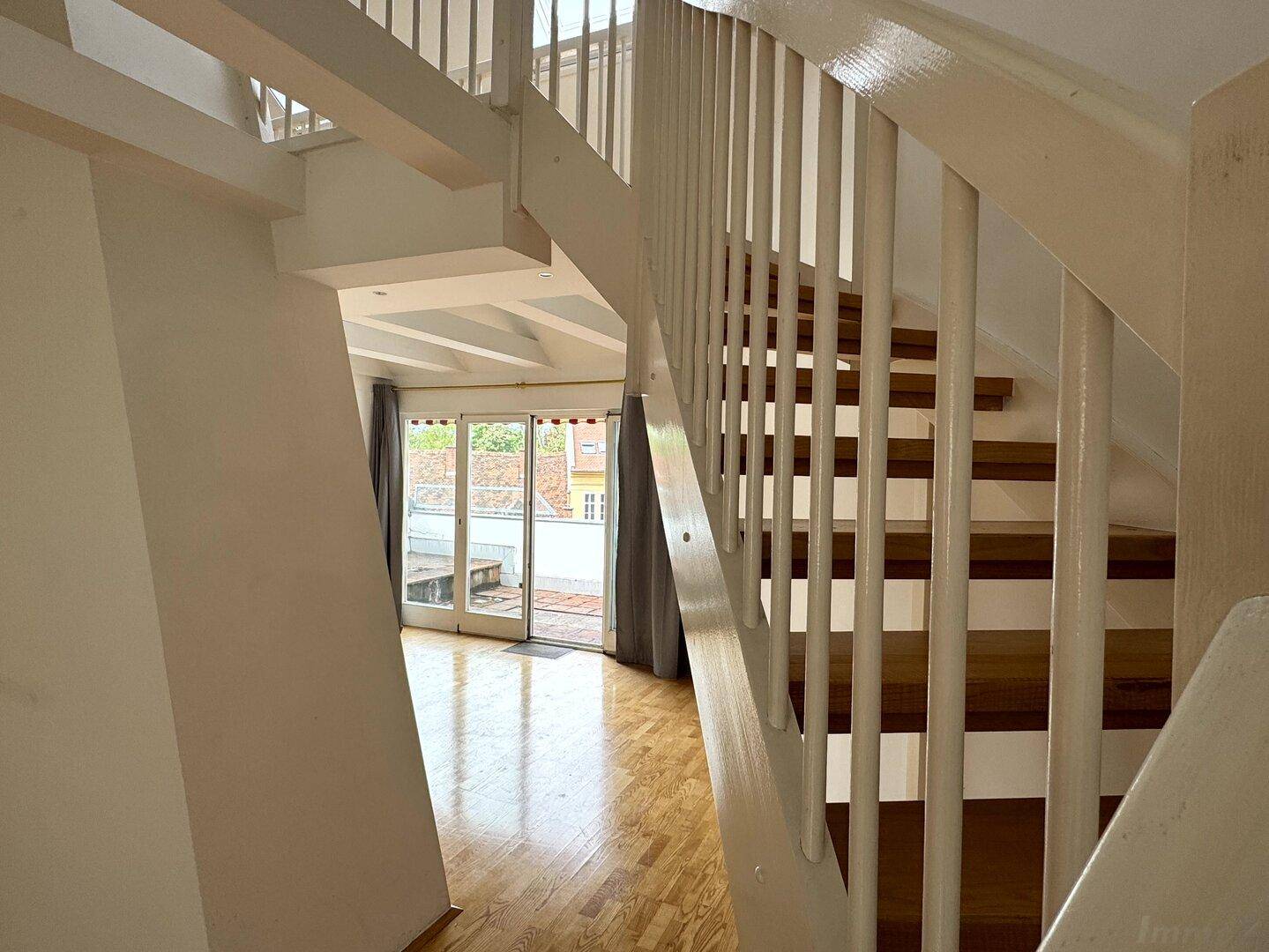 Blick von der Holztreppe auf den Wohnbereich mit Zugang zu einer sonnigen Terrasse durch eine Glasschiebetür.