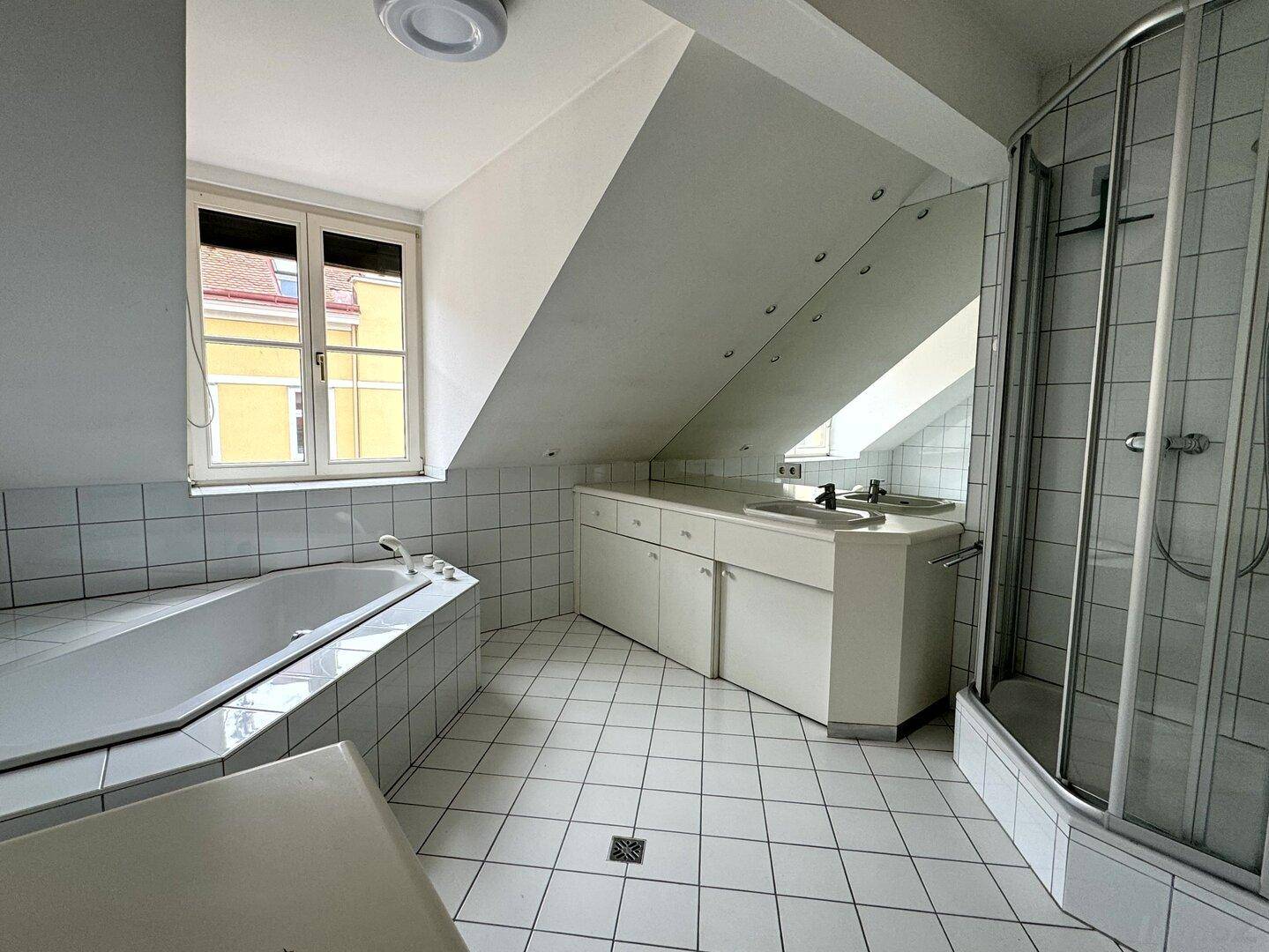 Helles Badezimmer mit weißen Fliesen, Eckbadewanne, Duschkabine, Doppelwaschbecken und Fenster.