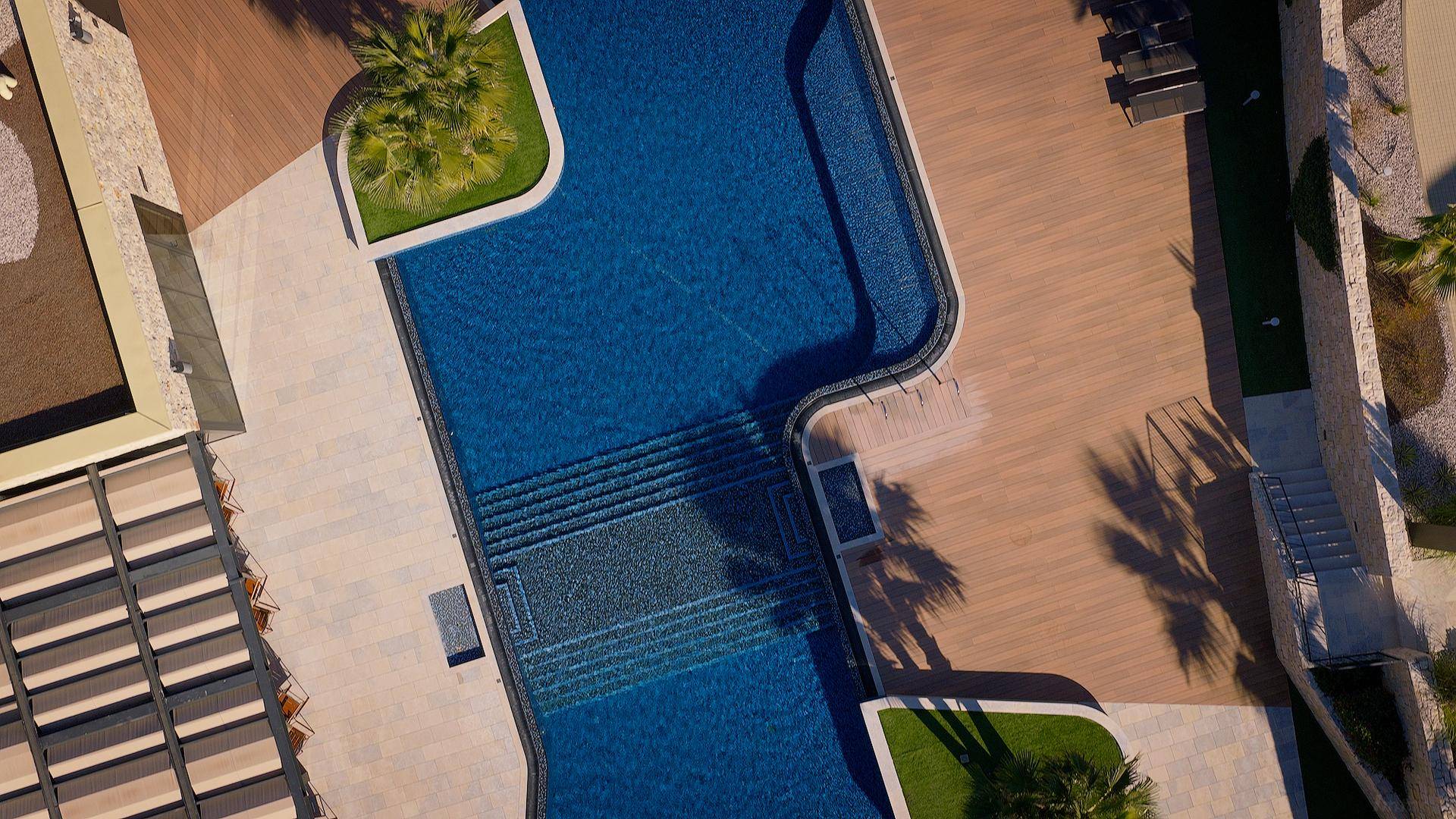 Vogelperspektive eines eleganten Pools mit Holzdeck und Liegebereichen, umgeben von Palmen.