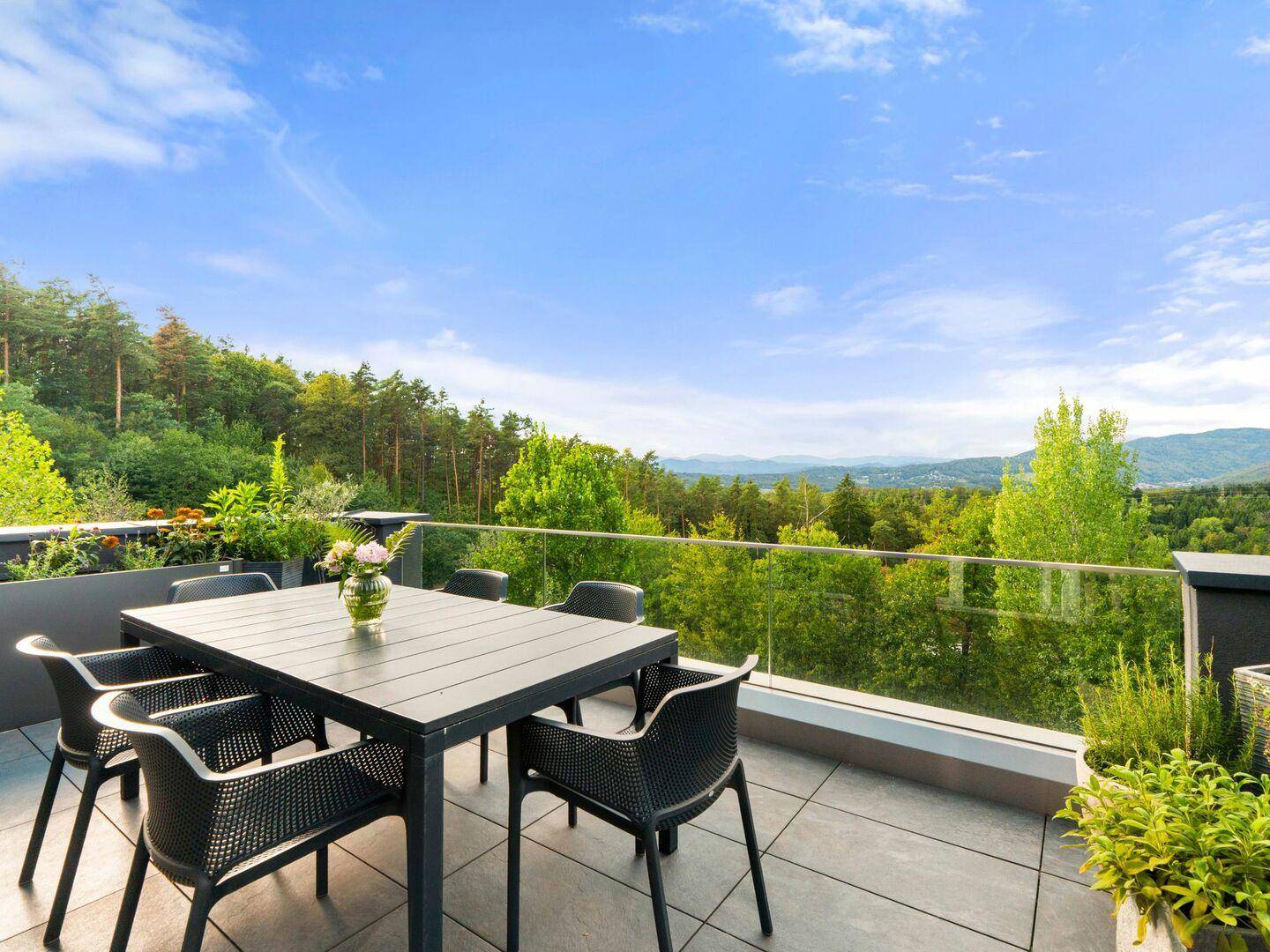 Große Dachterrasse mit Esstisch, Glasgeländer und weitem Blick über Wälder und Berge.