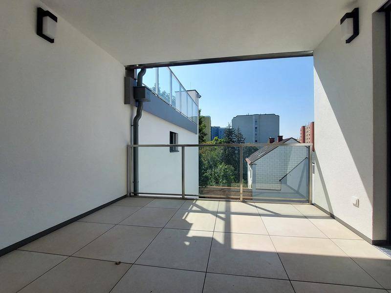 Überdachte Loggia mit Fliesenboden und Glasgeländer, bietet einen Blick ins Grüne.