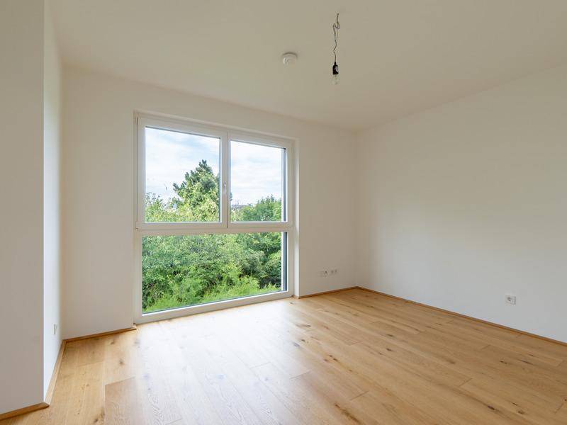 Leeres Schlafzimmer mit Holzboden und großem Fenster mit Blick ins Grüne.