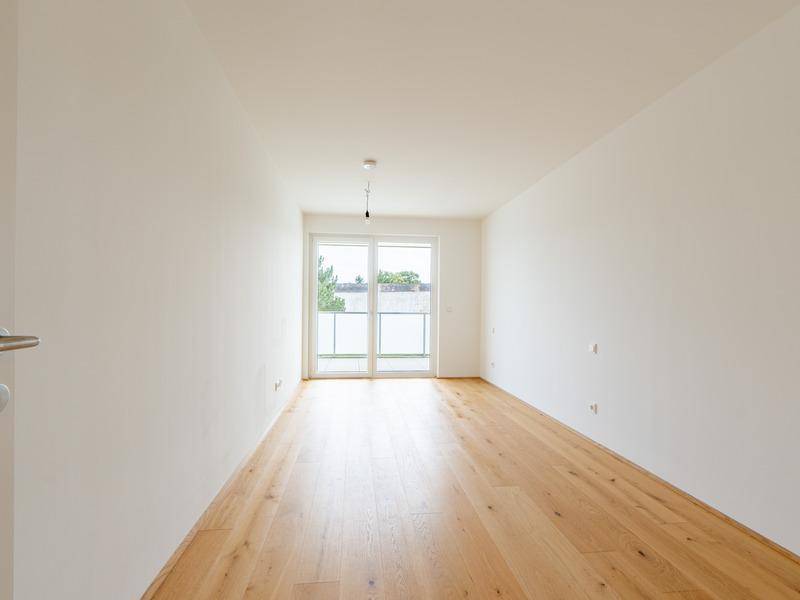 Helles, leeres Zimmer mit Holzboden und Zugang zu einem kleinen Balkon.