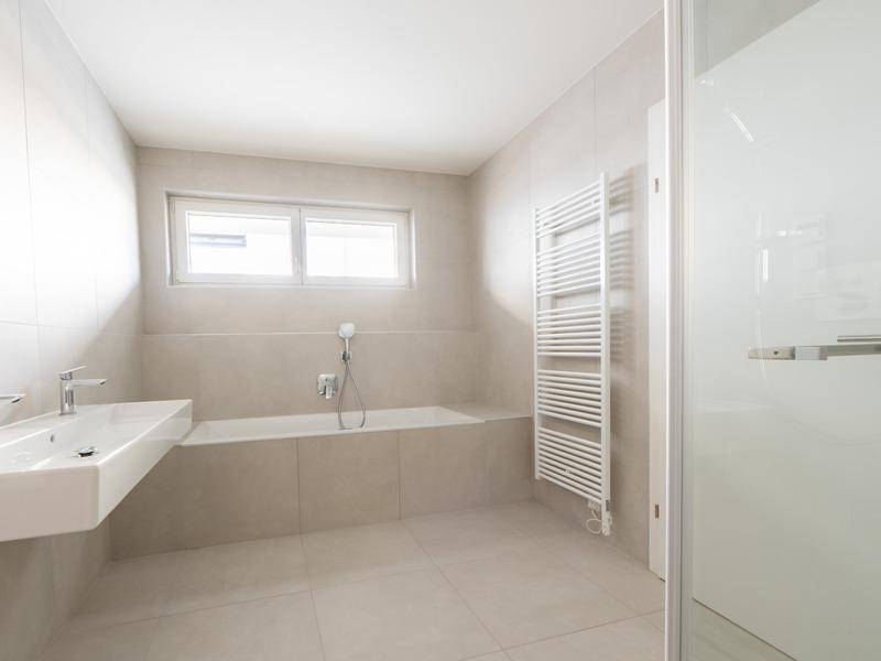 Modernes Badezimmer mit beigen Fliesen, Badewanne, Dusche, beheiztem Handtuchhalter und Fenster.