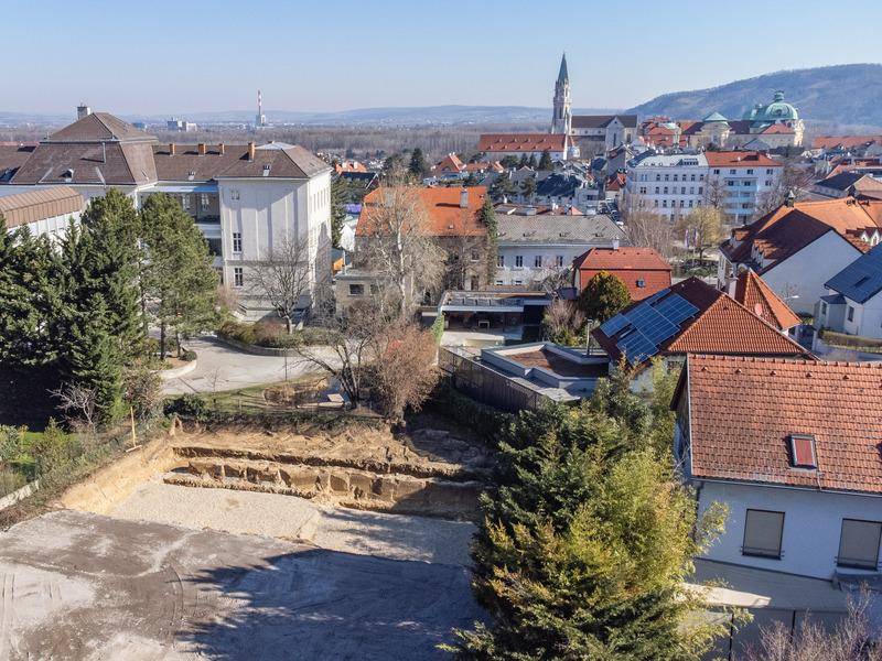 Luftaufnahme einer Baustelle mit ausgehobener Grube und Blick auf eine Stadt mit Kirche im Hintergrund.