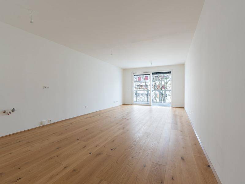 Helles, leeres Wohnzimmer mit neuem Holzparkettboden und Zugang zu einem Balkon.