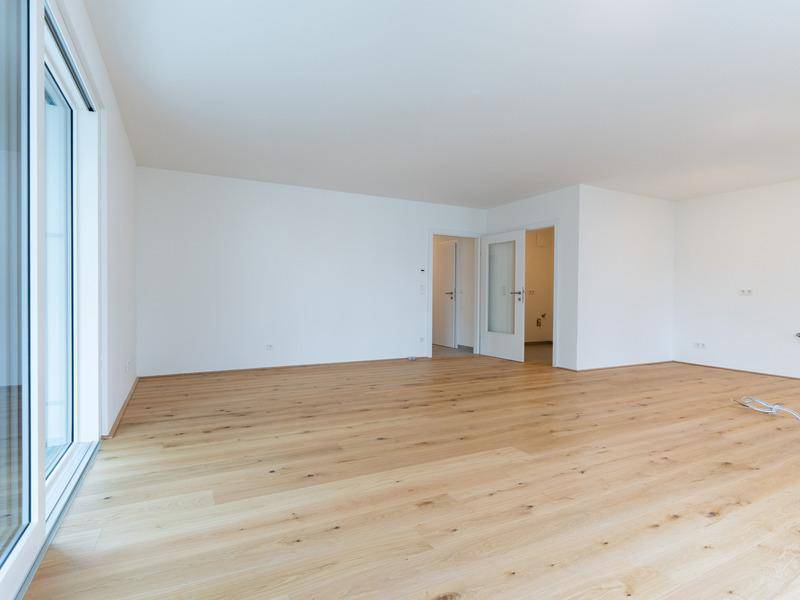 Geräumiger Wohnbereich mit hellem Holzboden, weißen Wänden und Zugang zu einem Außenbereich und weiteren Räumen.