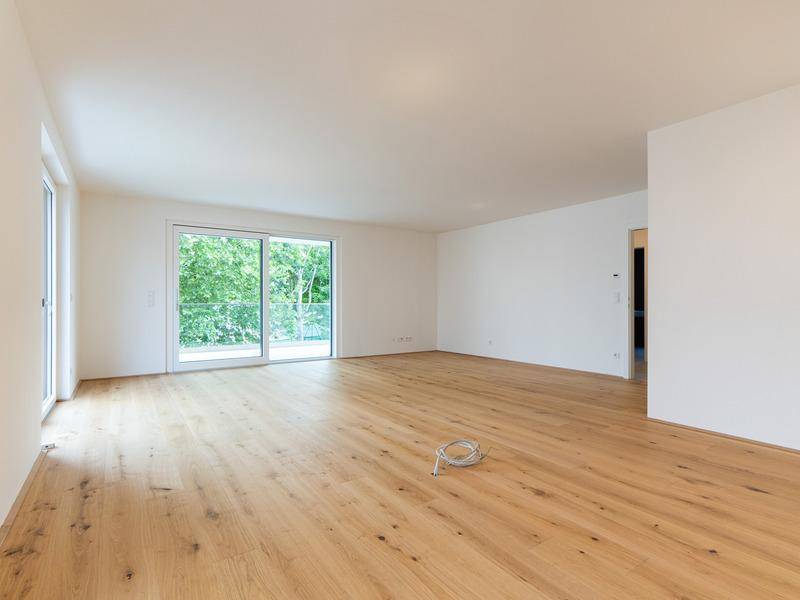 Lichtdurchfluteter Wohnbereich mit Holzboden und großer Schiebetür zu einem Balkon mit Grünblick.