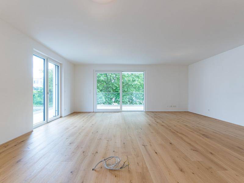 Großer, heller Wohnraum mit Holzboden und zwei Glastüren, die zu einem Balkon mit Grünblick führen.