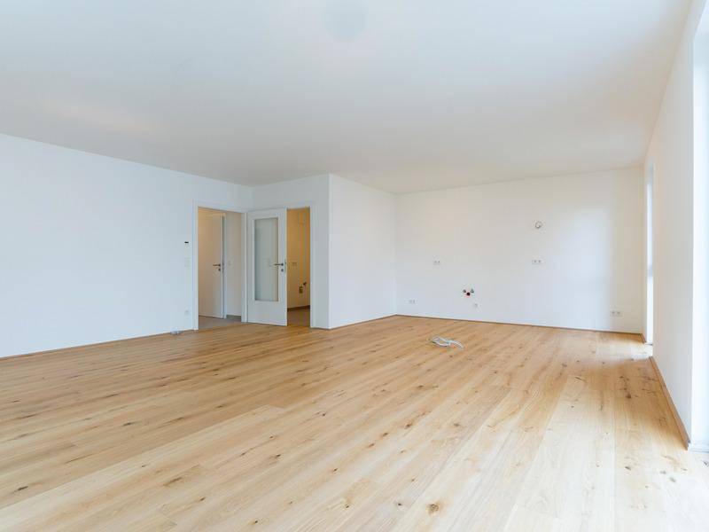 Geräumiger, leerer Raum mit hellem Holzfußboden, weißen Wänden und Zugang zu weiteren Zimmern.