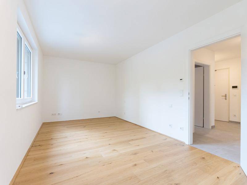 Heller, leerer Raum mit Holzboden, weißen Wänden und Fenster, angrenzend an einen gefliesten Flur.