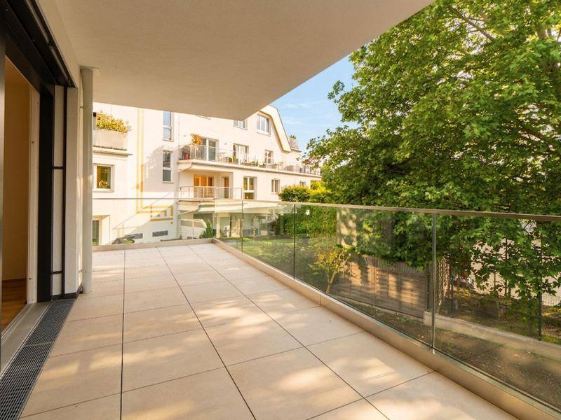 Großer, sonniger Balkon mit Fliesenboden, Glasgeländer und Blick auf Bäume und umliegende Gebäude.
