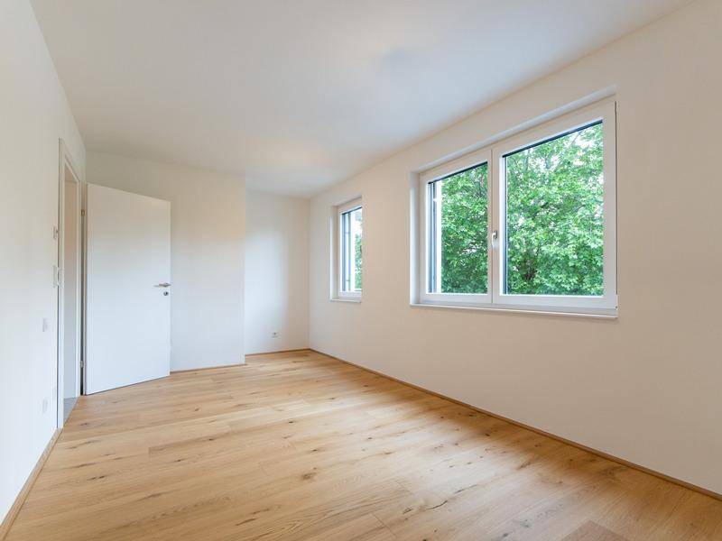Lichtdurchfluteter, leerer Raum mit Holzboden, weißen Wänden und Blick ins Grüne durch zwei Fenster.