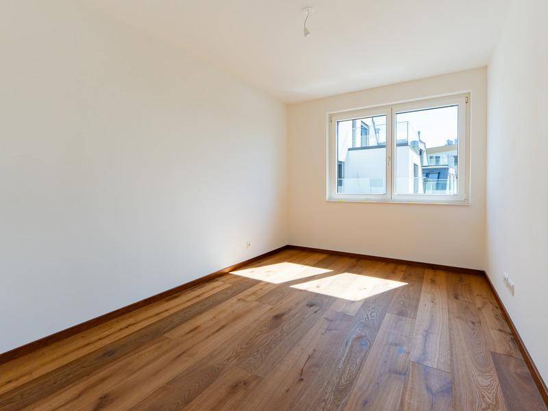 Leerer Raum mit Holzboden und einem großen Fenster, das viel Tageslicht hereinlässt.
