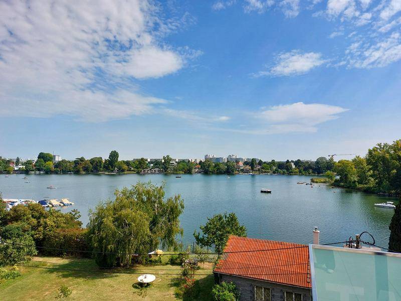 Weitläufiger Ausblick von einem Balkon über einen See mit bewaldeten Ufern und städtischer Skyline in der Ferne.