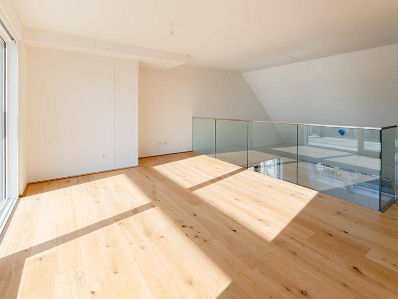 Heller, unmöblierter Raum mit Holzboden, großem Fenster und moderner Glasbrüstung.
