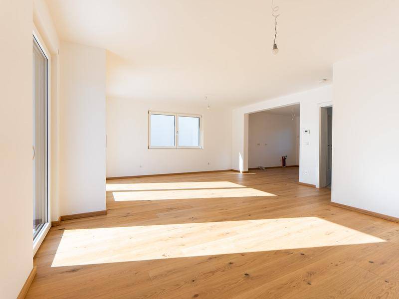 Heller, leerer Wohnraum mit Eichenparkettboden und großen Fenstern, die viel Licht hereinlassen.